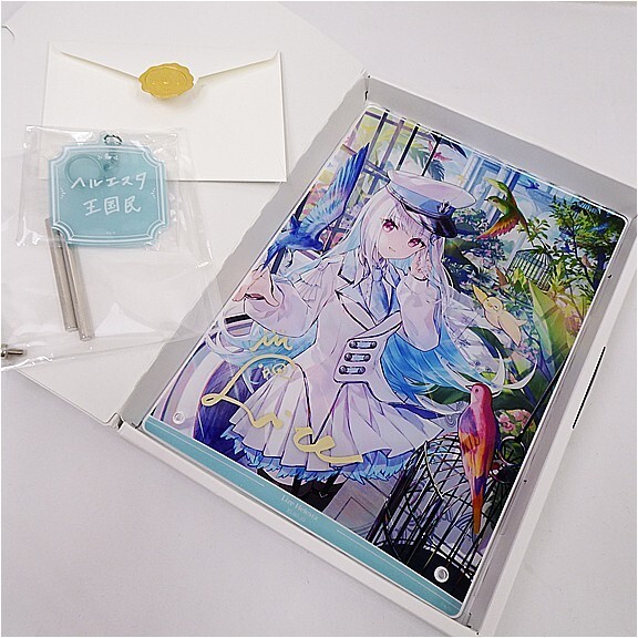 Open white box revealing a colorful anime artwork of a girl in a white outfit with blue hair, a bird nearby, inside a protective sleeve; envelope with a yellow wax seal sits beside and a blue-green tag in plastic adds context