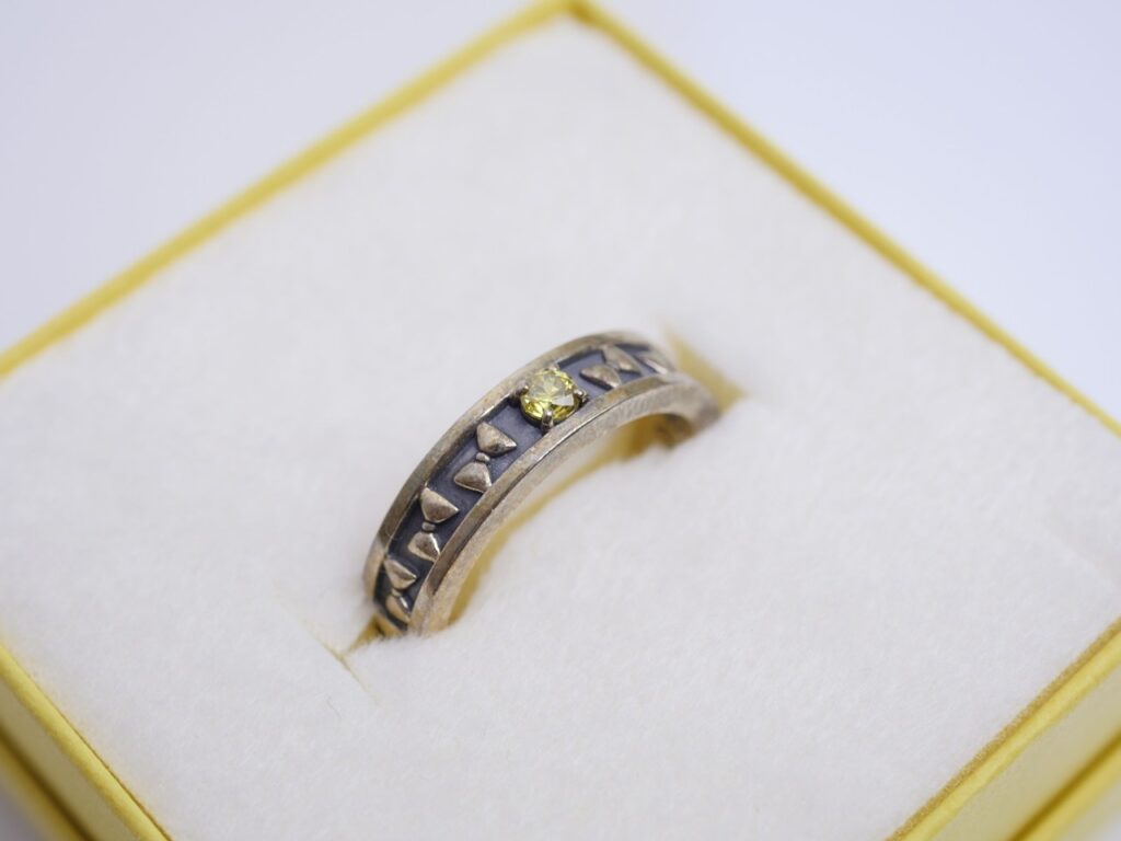 2460918 - Vtuberグッズ買取専門店Vtrader Silver-toned ring with a small yellow gemstone and chevron-shaped inlays, resting in a padded jewelry box.