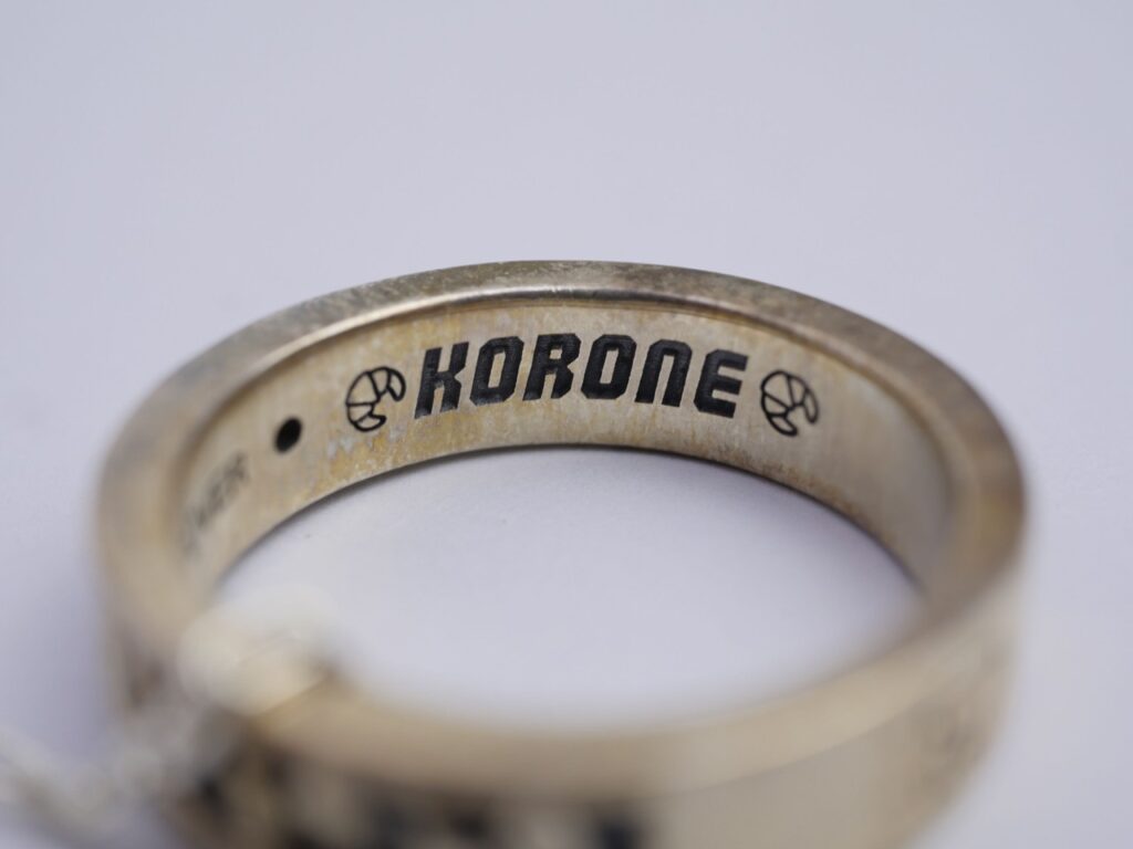 2460925 - Vtuberグッズ買取専門店Vtrader Close-up of a worn metal ring showing the inner engraving 'KORONE' with small circular logos on both sides.