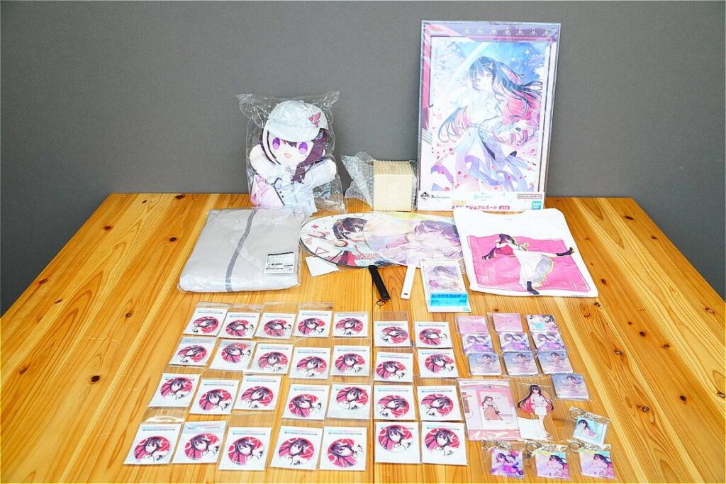 Collection of anime merch spread on a wooden table: plush doll, posters, towels, and trading cards featuring a pink-haired character.