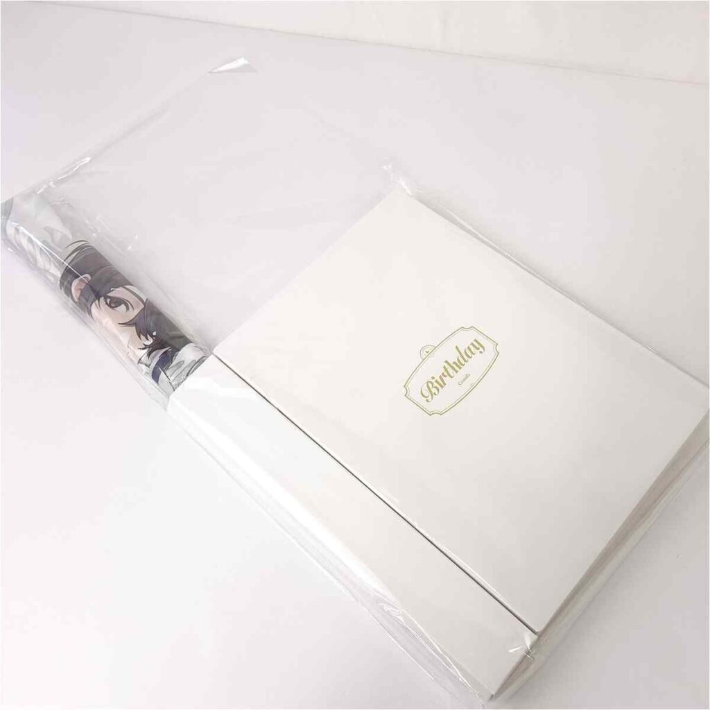 White gift box with a gold 'Birthday' label, wrapped in clear plastic, with a hint of anime artwork visible on the side.