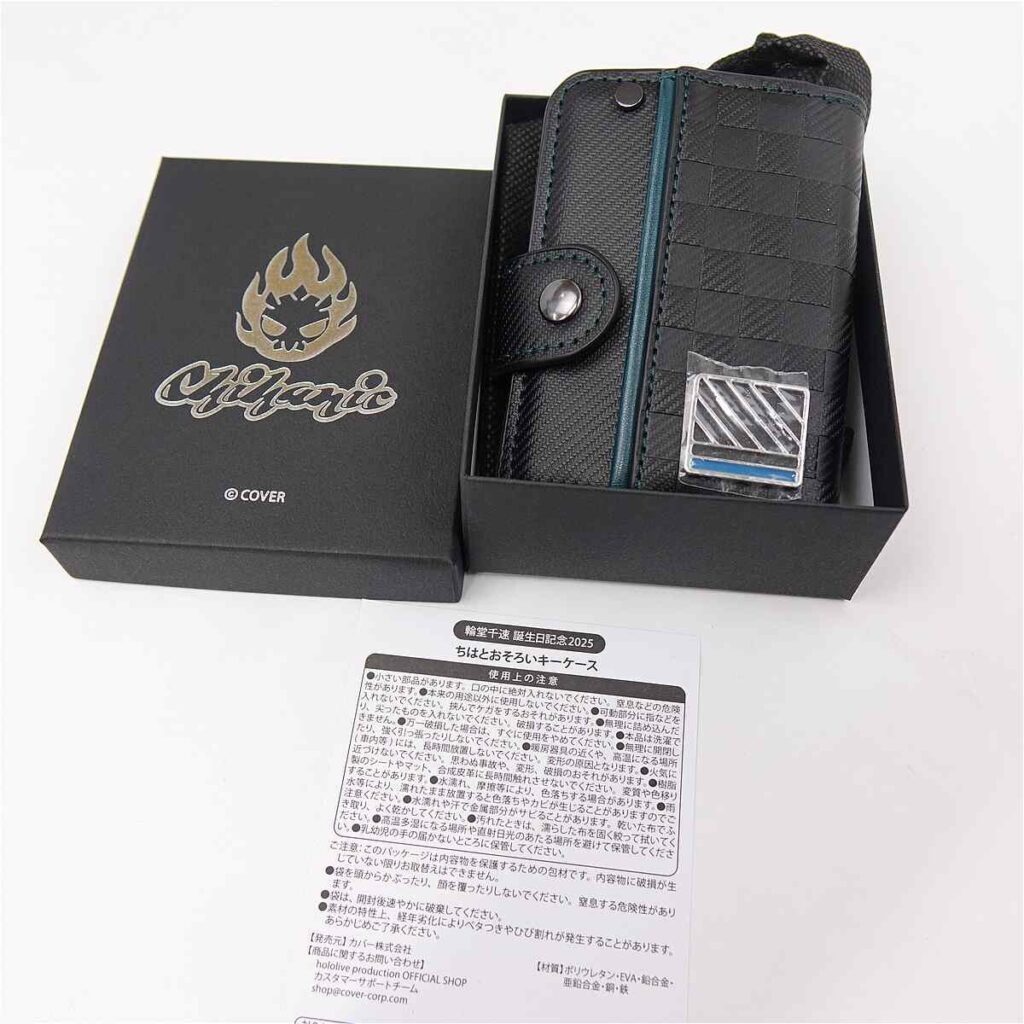 Black textured wallet/phone case in an open black gift box, with a teal zipper and metal badge. Short strap and snap on the side are visible in the case slot.