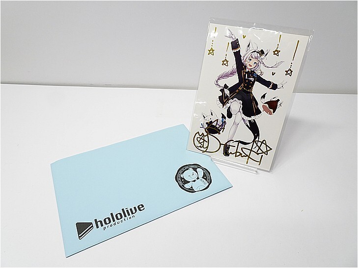 Light blue envelope with Hololive Production logo beside a laminated card featuring a stylized anime girl in a black outfit and gold stars.