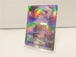 Holographic anime trading card on a clear stand featuring a cheerful orange-haired girl in a green outfit, with 'Rise from the ashes' text.