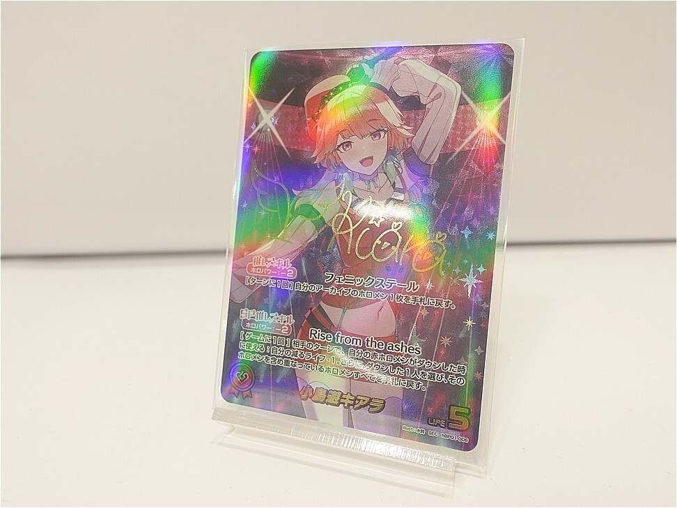 Holographic anime trading card on a clear stand featuring a cheerful orange-haired girl in a green outfit, with 'Rise from the ashes' text.