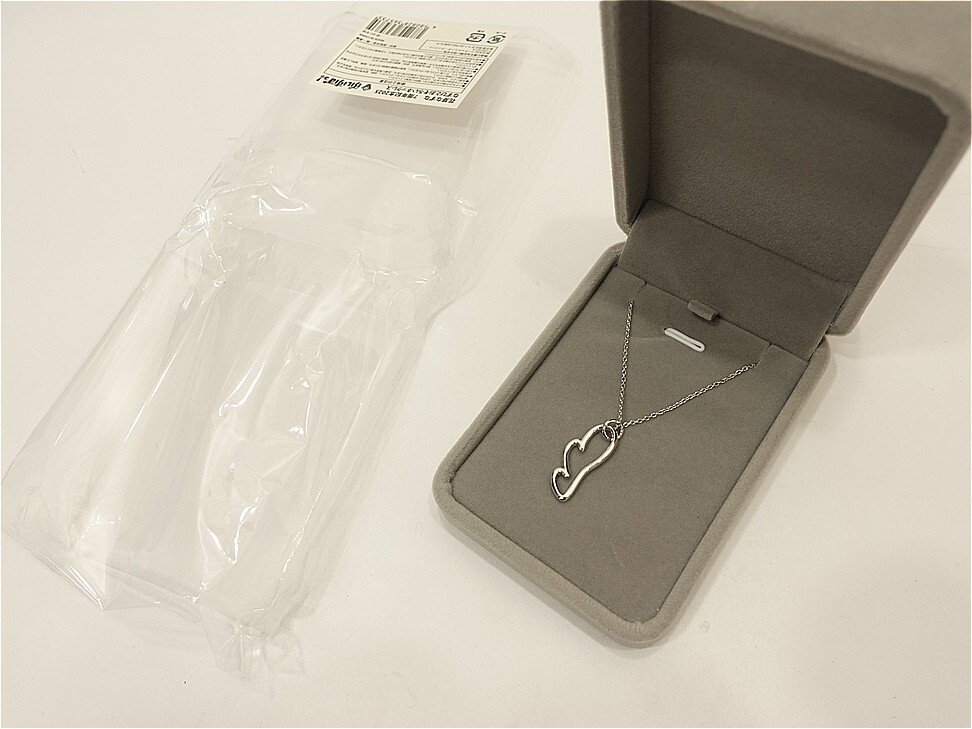Heart-shaped pendant necklace in an open jewelry box with a gray interior, placed on a white surface (plastic packaging nearby).