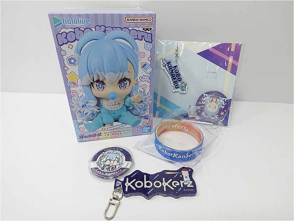 Blue-haired chibi figure box from Hololive with accompanying accessories laid out: a blue wristband, round pin, metal keychain, and a holographic-looking badge/sticker, all on a white surface.