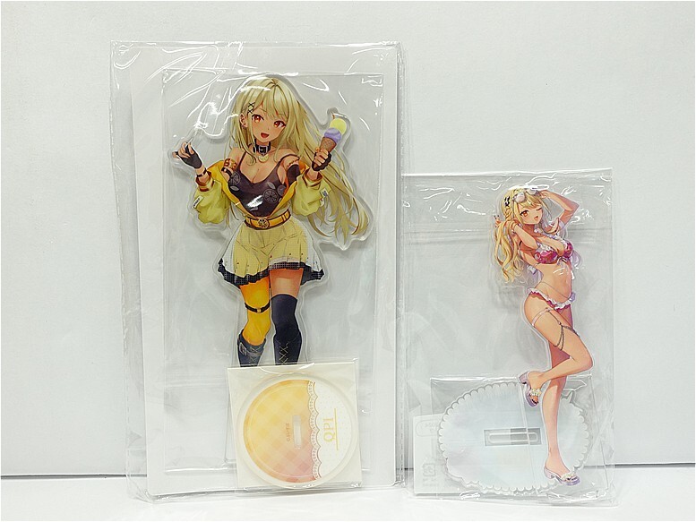 k12133642231 - Vtuberグッズ買取専門店Vtrader Two anime-style character standees in clear packaging: a larger yellow-outfit girl with microphone and a smaller pink bikini girl, plus accessories like a CD and a stand disc visible.