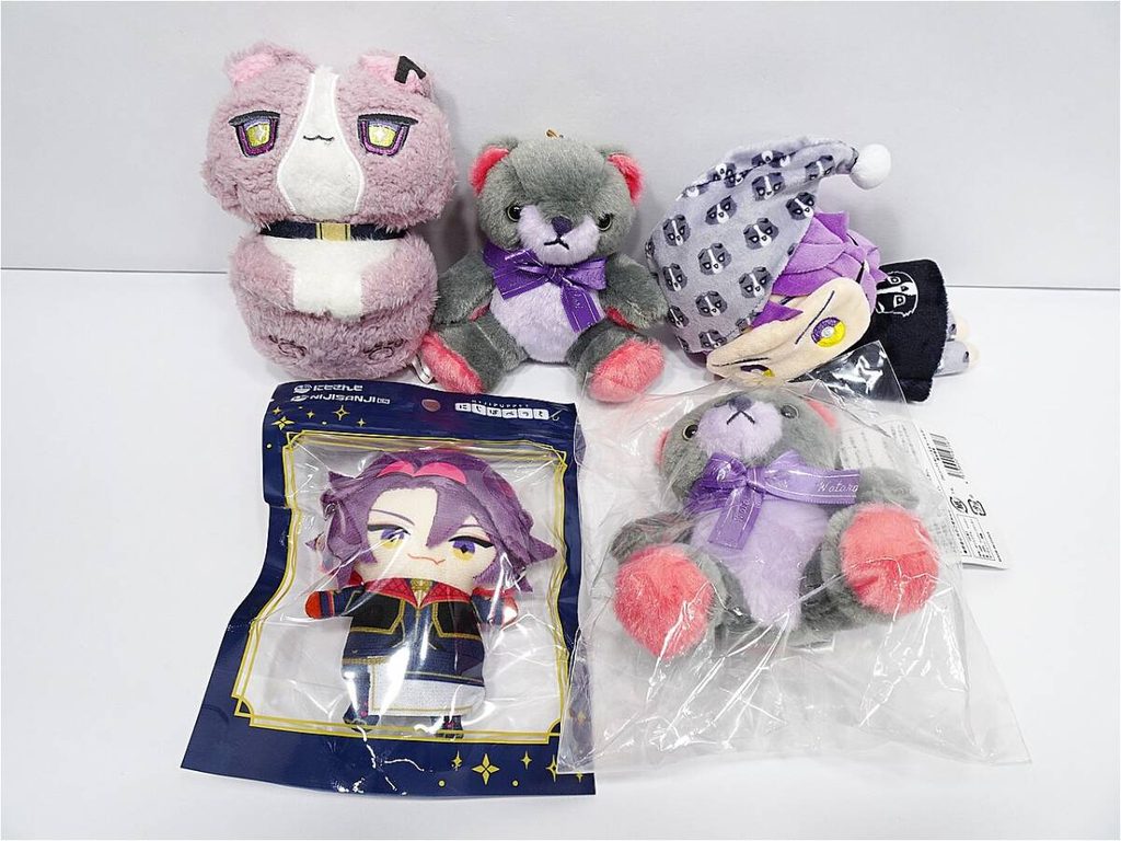 Collection of small plush toys: pink cat, gray bear with purple bow, lavender and gray hat, plus a boxed chibi character figure in dark blue packaging with gold border.